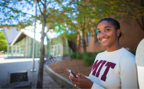 Photo: Young MIT student holds a phone in her hand, smiling and looking into the distance. She is wearing a white long sleeve with the letters MIT written in red across it. She stands in front of Kresge Auditorium, which is just out of focus in the background.