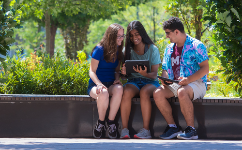 Photo: Three students sit on a stone bench on a sunny day. They are smiling and looking together at an iPad. They're each wearing shorts and sneakers with different color short sleeve shirts.